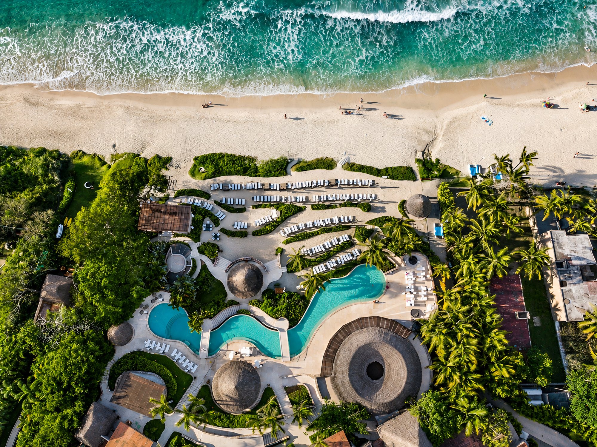 Vista cenital de Kupuri Beach Club: albercas, palapas y camastros junto a la playa y el oleaje